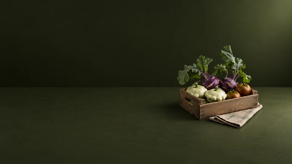 A wooden crate holding purple kohlrabi, pale pattypan squash and striped tomatoes beside folded linen.