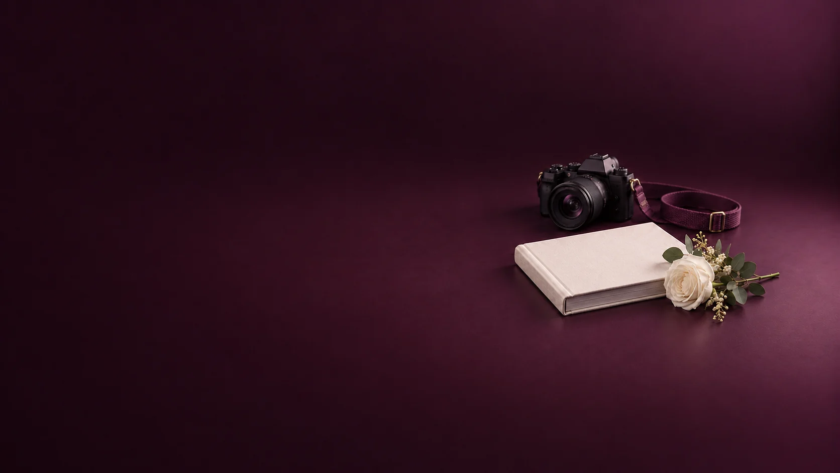 An unbranded camera with plum strap beside an ivory linen album and white rose.
