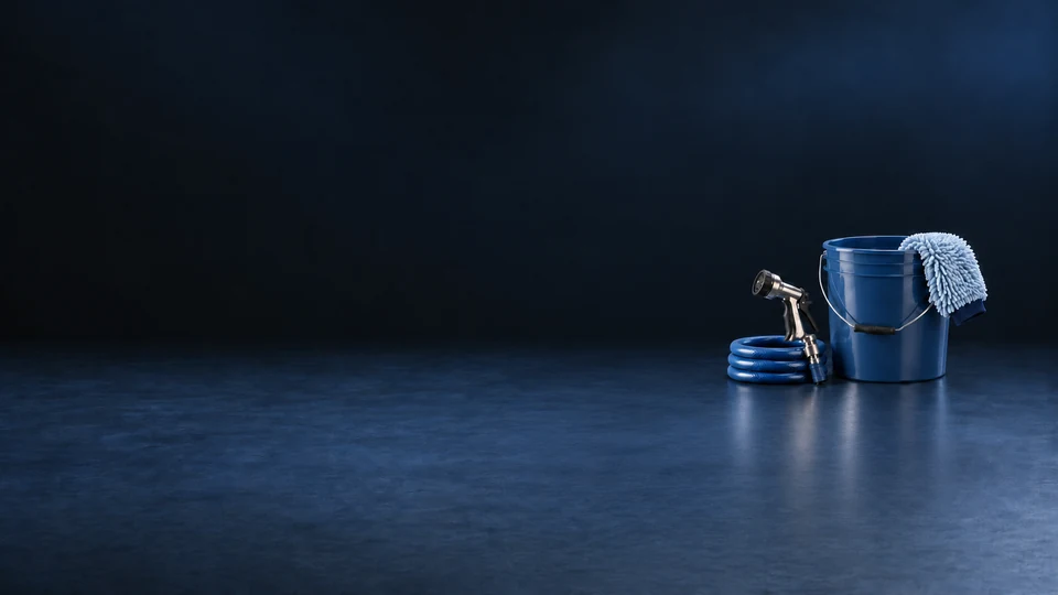 A blue wash bucket, pale mitt, metal nozzle and coiled blue hose on a navy workshop surface.