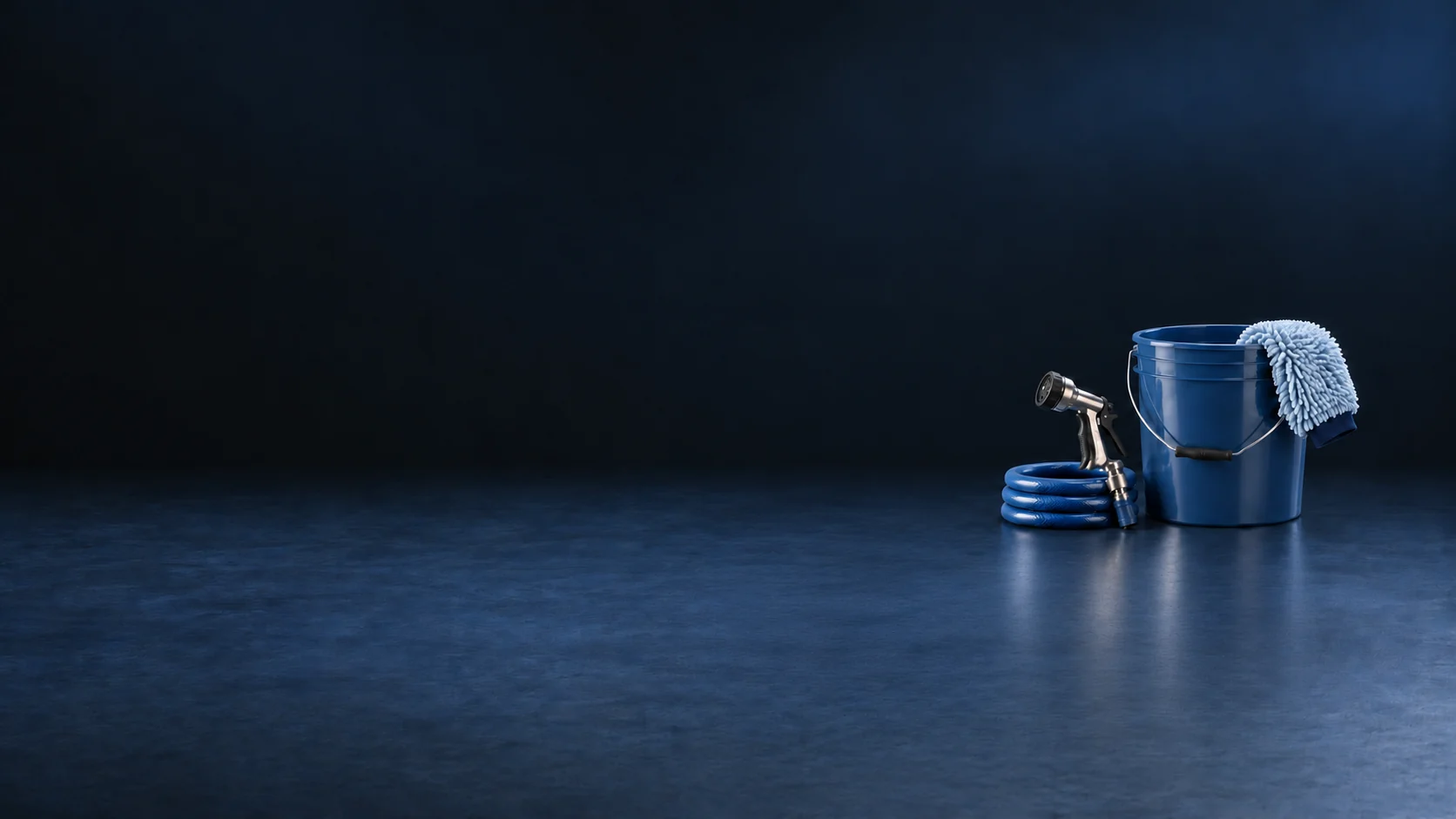 A blue wash bucket, pale mitt, metal nozzle and coiled blue hose on a navy workshop surface.