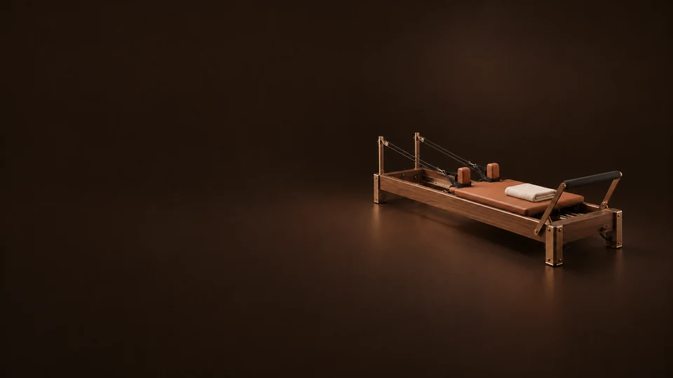 A walnut Pilates reformer with terracotta upholstery, bronze fittings and a folded ivory towel sits on a dark brown floor.