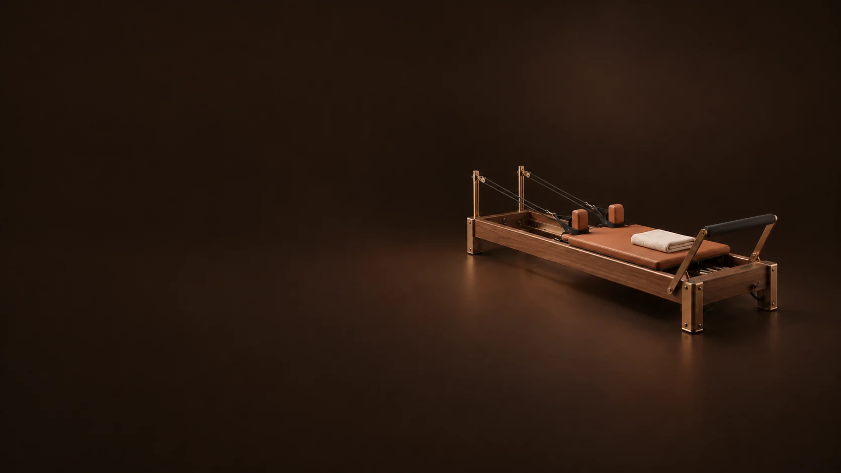 A walnut Pilates reformer with terracotta upholstery, bronze fittings and a folded ivory towel sits on a dark brown floor.