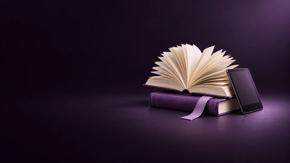 An open blank book with fanned paper leaves above a violet volume beside an e-reader and cloth bookmark.