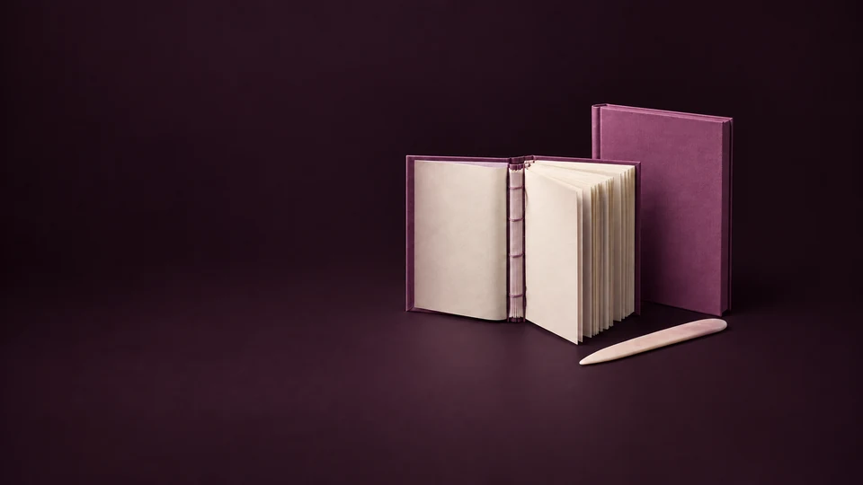 An open orchid casebound book exposes cream pages and a sewn spine beside a separate hardcover shell and bone folder.