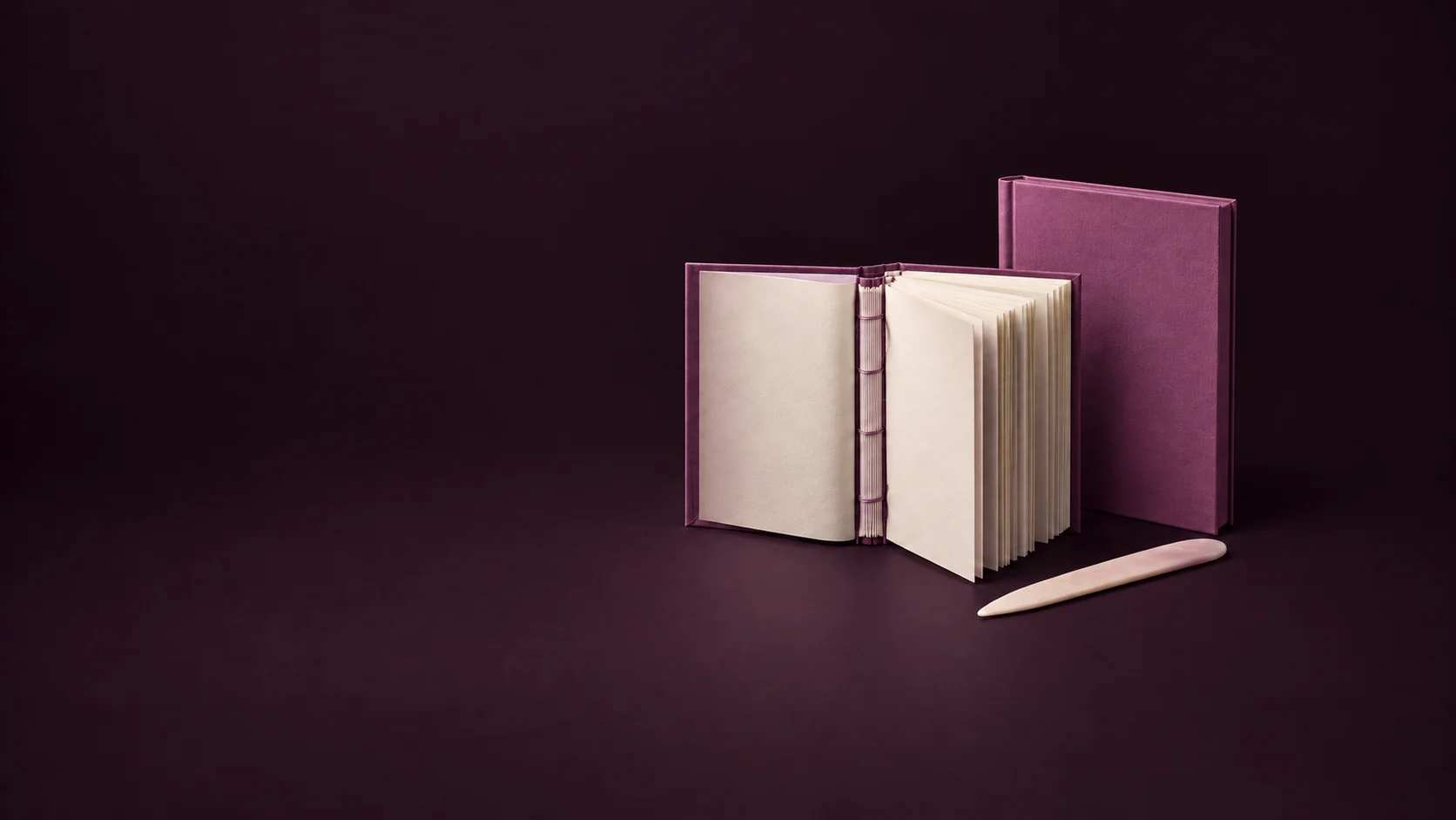 An open orchid casebound book exposes cream pages and a sewn spine beside a separate hardcover shell and bone folder.