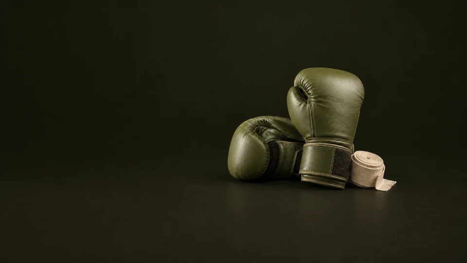 Olive boxing gloves rest with a rolled cream hand wrap on a dark studio floor.
