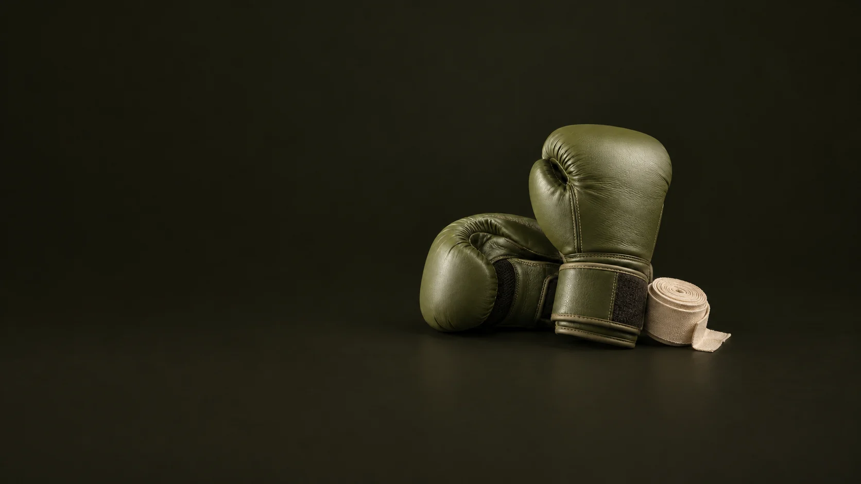 Olive boxing gloves rest with a rolled cream hand wrap on a dark studio floor.